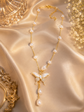 A gold chain necklace with scattered white pearls and a central butterfly pendant made of white mother-of-pearl and clear crystals, displayed on draped champagne satin with ornate gold props.