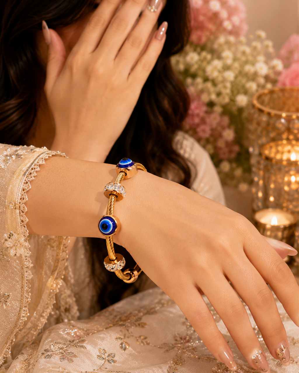 A gold-plated charm bracelet with blue evil eye beads and clear crystal accents, worn on a woman's wrist. The image features a warm, soft-focus background with pink flowers and a lit candle, and the woman is wearing a cream-colored embroidered dress.