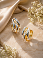A pair of gold and silver twisted hoop earrings displayed on a beige marble surface with draped champagne silk and small white flowers.