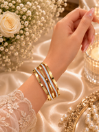 A close-up of a woman's hand and wrist wearing a stack of gold bangles, some embellished with clear crystals. The background features elegant cream satin fabric, white roses, and a lit candle in a glass holder.