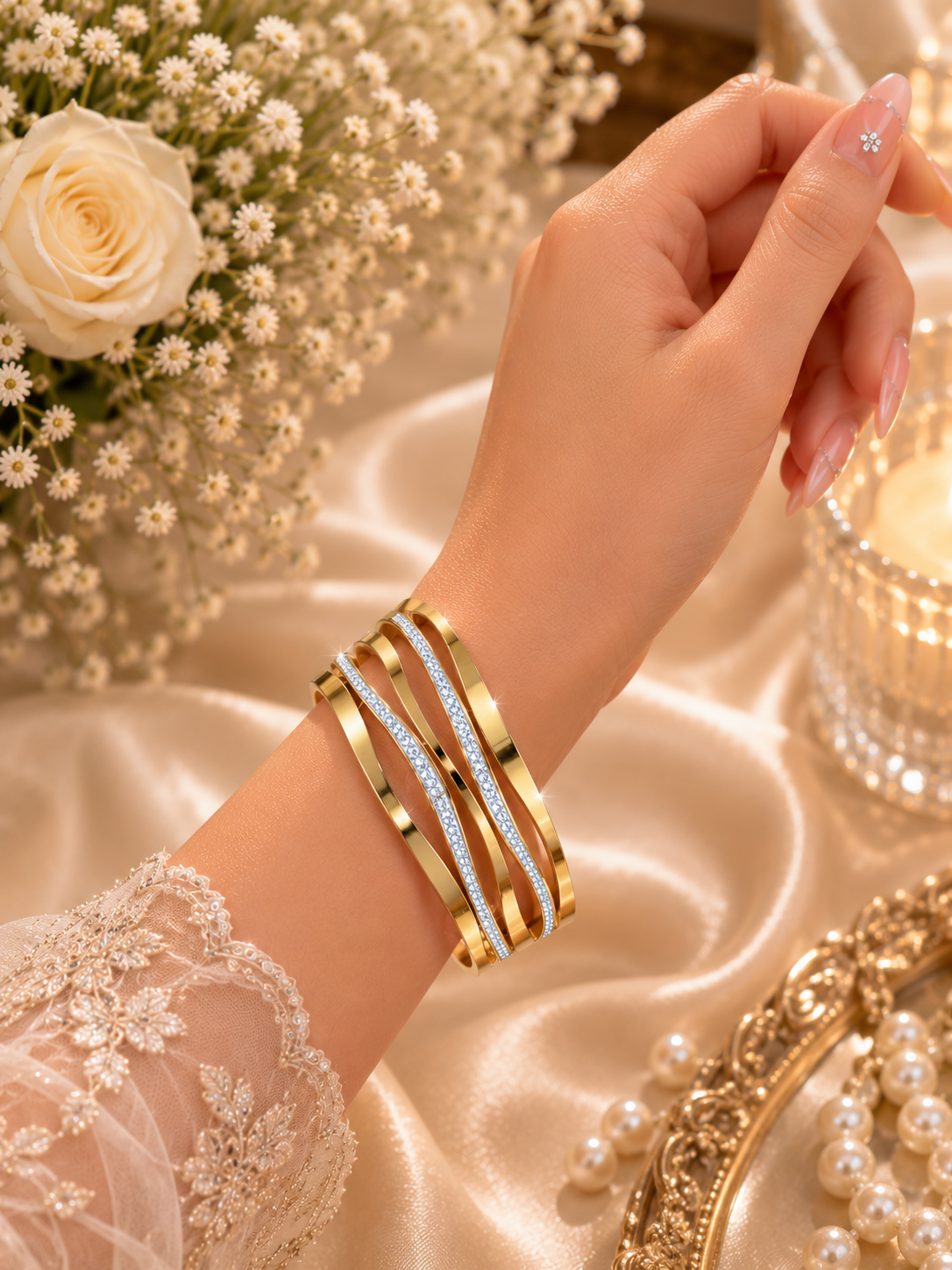 A close-up of a woman's hand and wrist wearing a stack of gold bangles, some embellished with clear crystals. The background features elegant cream satin fabric, white roses, and a lit candle in a glass holder.