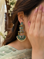 A close-up of a woman wearing a large, ornate gold-toned statement earring with green stones and white pearl-like accents.