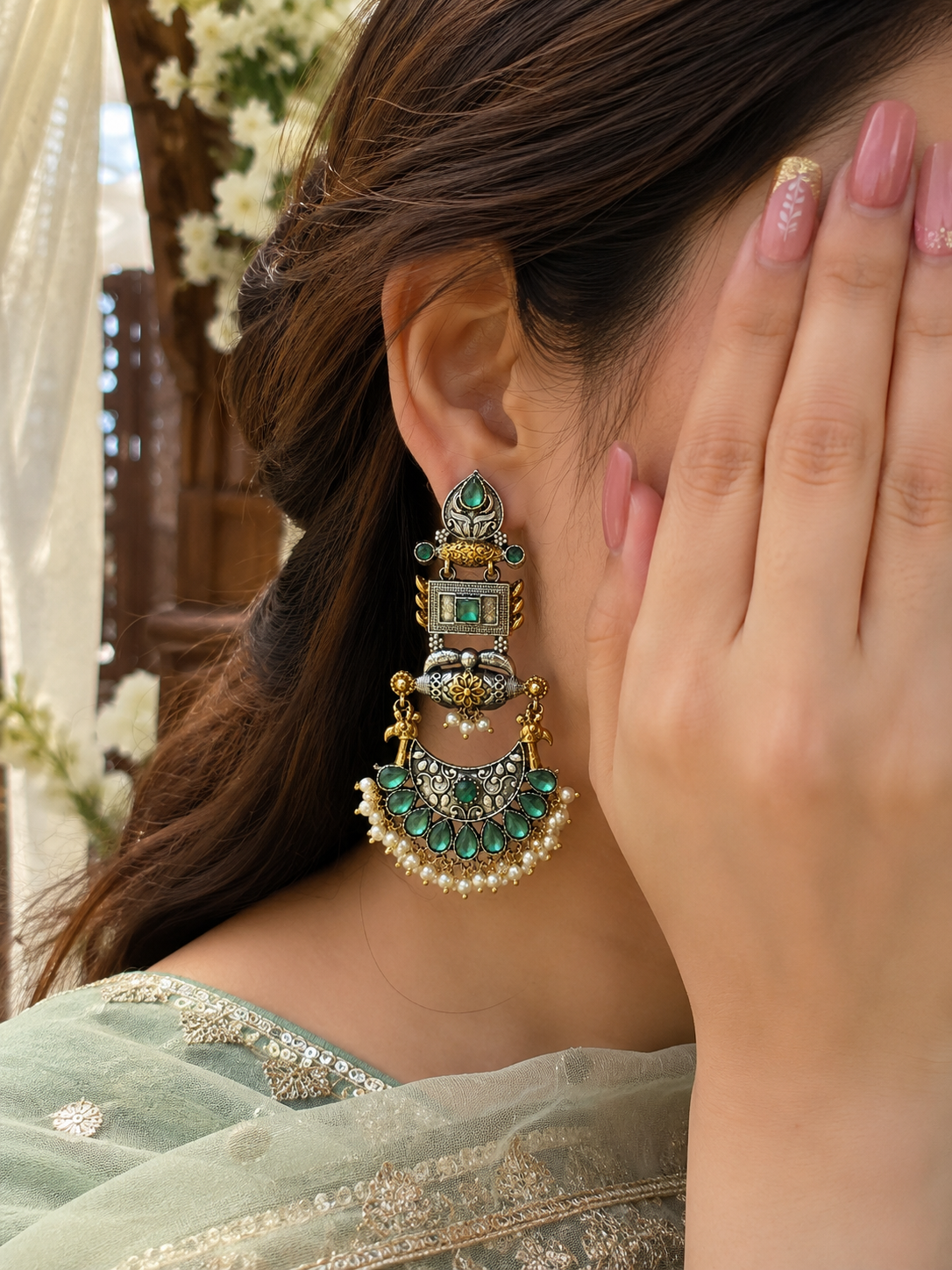 A close-up of a woman wearing a large, ornate gold-toned statement earring with green stones and white pearl-like accents.