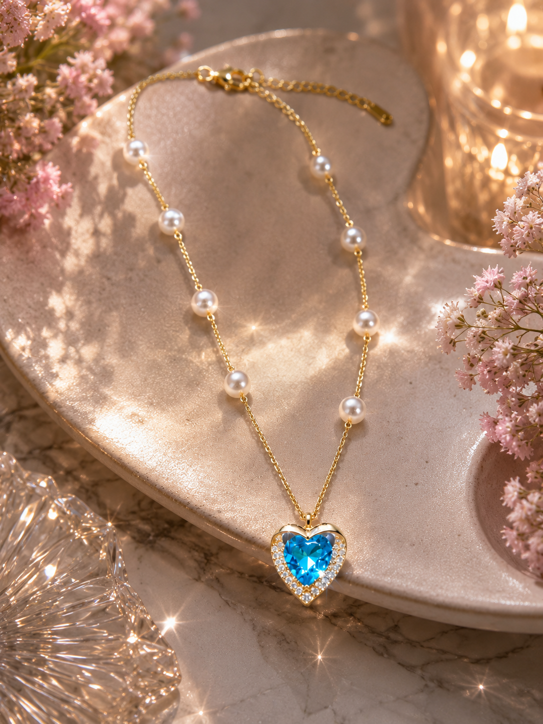 A gold necklace with a heart-shaped blue gemstone pendant, accented with small white pearls along the chain, displayed on a beige ceramic dish with pink flowers and warm bokeh lights.