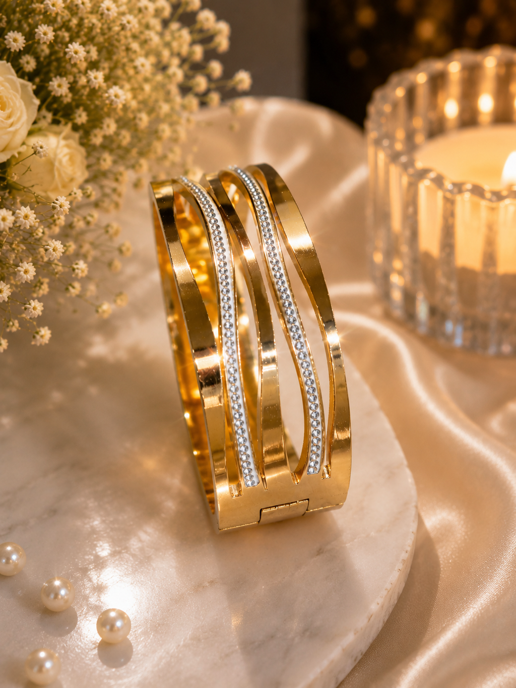 A gold-toned wide cuff bracelet with two inlaid rows of small clear stones, displayed on a marble slab with satin fabric, pearls, flowers, and a lit candle in the background.
