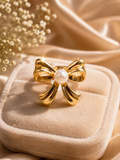 A gold bow-shaped ring with a white pearl at the center, displayed on a beige velvet jewelry box with soft satin fabric and dried flowers in the background.