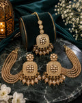 Gold-toned Indian bridal jewelry set featuring a maang tikka, earrings, and a necklace displayed on a black velvet stand, arranged on a dark marble surface with decorative flowers and a lantern.