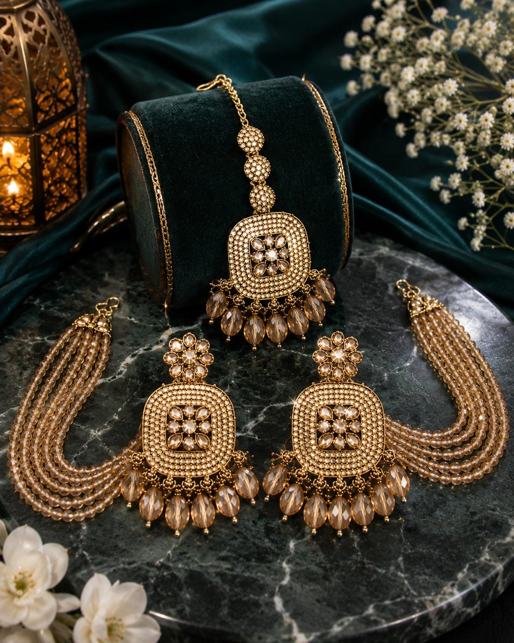 Gold-toned Indian bridal jewelry set featuring a maang tikka, earrings, and a necklace displayed on a black velvet stand, arranged on a dark marble surface with decorative flowers and a lantern.
