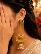 A close-up lifestyle shot of a woman wearing an ornate gold-toned traditional Indian jhumka earring with pink, green, and white stone accents and dangling chains.
