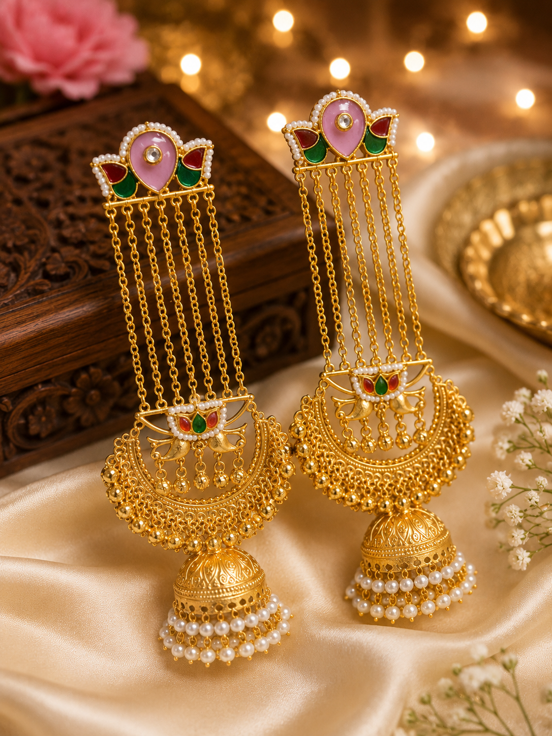 A pair of ornate gold-toned traditional Indian jhumka earrings with pink, green, and white enamel details.